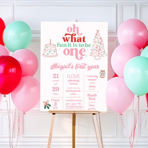 May include: A first birthday sign in pink and white, with the words "Oh what fun it is to be one." The sign features a cake, Christmas tree, and a list of facts about the child. Pink, red, and teal balloons are arranged around the sign.