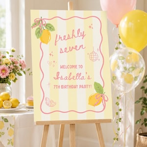 May include: A party sign on an easel with a yellow and white striped background. The sign reads "freshly seven" and "Welcome to Isabella's 7th Birthday Party!" with lemon illustrations and pink accents. Balloons in pink, yellow, and clear with lemon designs are in the background.
