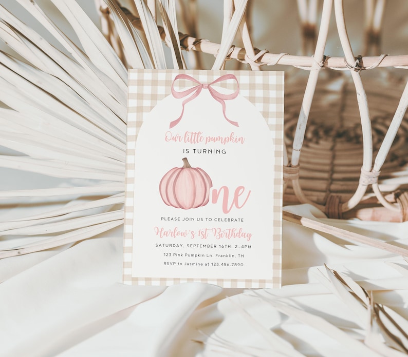 May include: A light pink and tan gingham patterned invitation card with a pink bow and pumpkin illustration. The text reads "Our little pumpkin is turning one." Additional text includes the event details: date, time, location, and RSVP information.