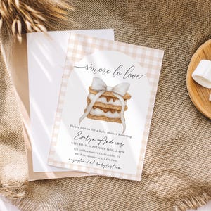 Könnte beinhalten: Eine Babyparty-Einladung mit einer Illustration eines S'mores, gebunden mit einer weißen Schleife. Die Einladung hat einen beigen Vichy-Rand und enthält den Text "smore to love" sowie Eventdetails für Evelyn Andrews.