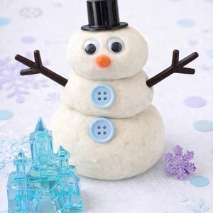 Pu&ograve; includere: Un pupazzo di neve stravagante fatto di pasta, con un cappello a cilindro nero, occhi finti e un naso arancione. Braccia di ramoscelli marroni e dettagli di bottoni blu completano il design. Sono presenti anche un castello blu e un fiocco di neve viola.