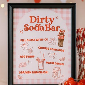 May include: A framed sign with the text "Dirty Soda Bar" in red, with instructions and illustrations for making a soda. The background is a pink and white gingham pattern. Red and white striped straws and cherries are in the foreground.