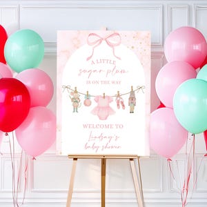 May include: A baby shower welcome sign on an easel, featuring a pink and gold marble background. The sign reads "A Little Sugar Plum Is On The Way" with illustrations of a mouse, tutu, ballet shoes, and nutcracker. Pink, red, and mint balloons surround the sign.