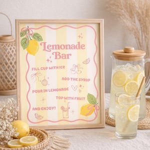 K&ouml;nnte beinhalten: Ein gerahmtes Schild mit den Worten "Lemonade Bar" in rosa Schrift, mit Illustrationen von Zitronen und Anweisungen. Eine Glaskaraffe und ein Glas Limonade mit Zitronenscheiben sind ebenfalls zu sehen, zusammen mit einem geflochtenen Tablett mit Zitronenscheiben.