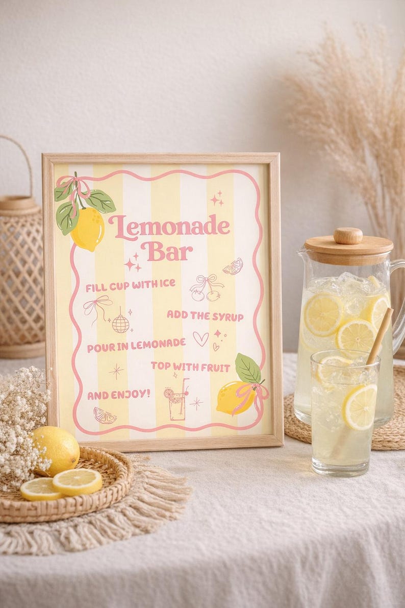 K&ouml;nnte beinhalten: Ein gerahmtes Schild mit den Worten "Lemonade Bar" in rosa Schrift, mit Illustrationen von Zitronen und Anweisungen. Eine Glaskaraffe und ein Glas Limonade mit Zitronenscheiben sind ebenfalls zu sehen, zusammen mit einem geflochtenen Tablett mit Zitronenscheiben.