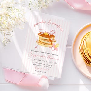 May include: A baby shower invitation with a pink and white striped design. The invitation features the text "pancakes & pacifiers" and a watercolour illustration of pancakes and a dummy. The invitation also includes event details.