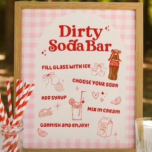 May include: A framed sign with a pink gingham pattern and the words "Dirty Soda Bar" in red script. The sign features illustrations and instructions for making a soda, plus a glass jar of red and white striped straws.
