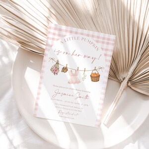 Peut inclure: Une invitation &agrave; une baby shower avec une bordure rose &agrave; carreaux et le texte "A Little Pumpkin is on her way!". L'invitation pr&eacute;sente des illustrations de v&ecirc;tements de b&eacute;b&eacute;, de fleurs et d'un panier de citrouilles. Le nom "Jasmine Smith" est &eacute;galement affich&eacute;.