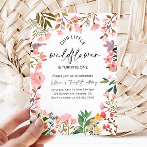 Può includere: Un invito di compleanno floreale con il testo "Our Little Wildflower is Turning One". L'invito presenta un bordo floreale ad acquerello con fiori rosa, viola e gialli e foglie verdi. L'invito è tenuto in mano.
