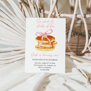 Puede incluir: Una tarjeta de invitación blanca con el texto "So sweet & stacks of fun" y "Lilah is turning one!" presenta una ilustración de acuarela de panqueques con un lazo rosa. La tarjeta incluye detalles del evento: fecha, hora, dirección e información de RSVP.