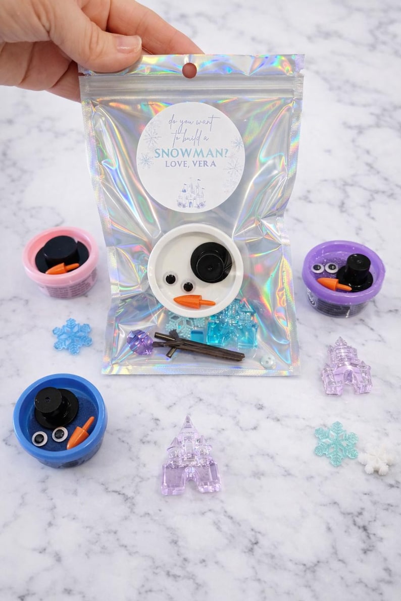 Pu&ograve; includere: Una busta trasparente con un kit per costruire un pupazzo di neve. Il kit include un cerchio bianco con parti del viso del pupazzo di neve, piccoli castelli di plastica, fiocchi di neve e contenitori con accessori per pupazzi di neve. La busta ha il testo "Do you want to build a snowman? Love, Vera."