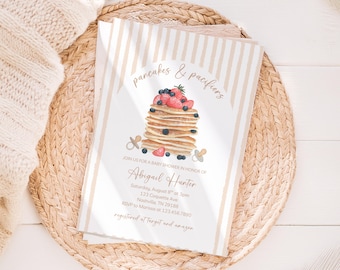May include: Baby shower invitation with a stack of pancakes topped with blueberries and strawberries. The invitation has the text "pancakes & pacifiers" and event details for Abigail Hunter. The invitation is on a beige striped background.