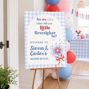 May include: A rectangular gender reveal sign on a wooden easel featuring a light blue gingham border. The text reads: he or she what will our little firecracker be? WELCOME TO Sierra & Carter's GENDER REVEAL, with a red and blue firework graphic.