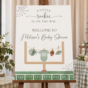 Op de afbeelding: Een wit rechthoekig bord met de tekst A LITTLE rookie IS ON THE WAY, WELCOME TO Melissa's Baby Shower. Het ontwerp bevat een football-doelpaal, een waslijn met een groen rompertje, een football, een helm en kleine schoentjes.