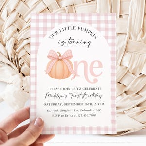 May include: A rectangular invitation card featuring a light pink and white gingham border. The centre displays a peach-coloured pumpkin with a matching gingham bow, the text "OUR LITTLE PUMPKIN is turning one," and event details for Madelyn's birthday.