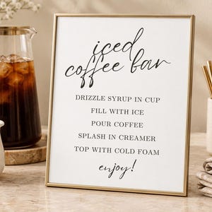 Könnte beinhalten: Ein goldgerahmtes Schild mit schwarzem Text: iced coffee bar, DRIZZLE SYRUP IN CUP, FILL WITH ICE, POUR COFFEE, SPLASH IN CREAMER, TOP WITH COLD FOAM, enjoy!. Das Schild steht auf einer Oberfläche neben einer Glaskaraffe mit Eiskaffee.