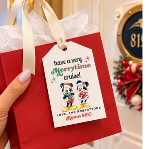 May include: A red gift bag with cream-colored ribbon handles and white tissue paper, featuring a white rectangular tag printed with the text "have a very Merrytime cruise!" and "LOVE, THE ROBERTSONS Room 8195" alongside two cartoon mice.