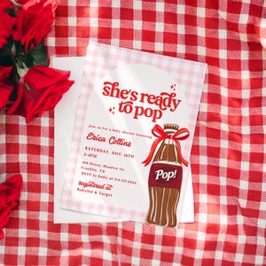 May include: A baby shower invitation with the text "She's ready to pop" in red script. The invitation features a cartoon soda bottle with a red bow. The invitation is on a white background with a red and white checkered pattern.