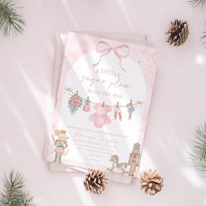 May include: A baby shower invitation with a pink and gold marble design. The invitation features the text "A little sugar plum is on the way" and illustrations of baby clothes and ballet shoes. The invitation is decorated with pinecones and greenery.