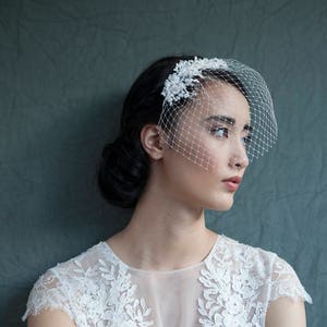 Puede incluir: Una novia con un vestido blanco de encaje con velo y una pieza floral para el cabello. La novia est&aacute; de pie frente a un fondo verde.