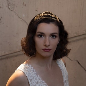 Può includere: Una donna con capelli castano scuro che indossa un top di pizzo bianco e una fascia per capelli con foglie dorate.