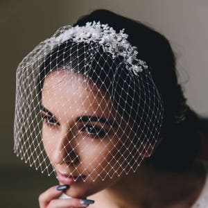 Puede incluir: Un velo de novia blanco con un delicado dise&ntilde;o floral y un patr&oacute;n de red. El velo lo lleva una persona con cabello oscuro y cubre la cara.