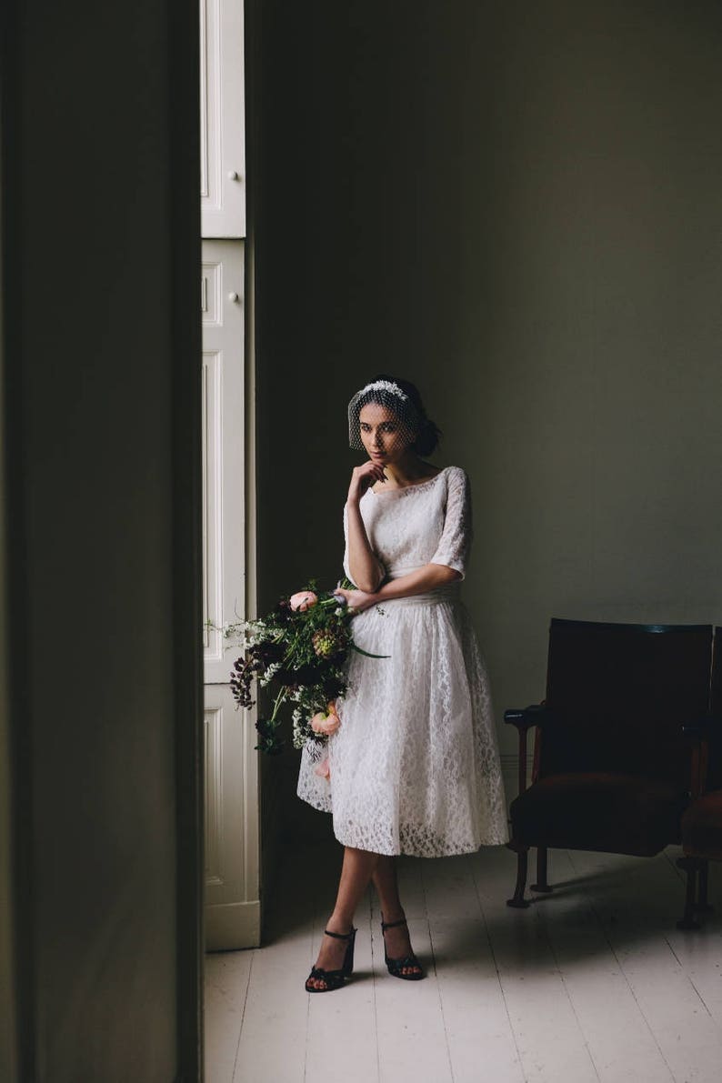Puede incluir: Una novia con un vestido blanco de encaje, velo y un ramo de flores. Est&aacute; de pie en una habitaci&oacute;n con una pared blanca y una puerta de madera.