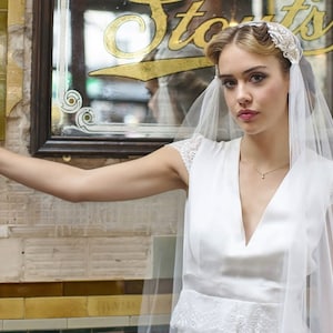 Puede incluir: Una mujer con un vestido blanco con un corpiño de encaje y un velo delicado con una diadema floral. Está de pie frente a una tienda vintage con un letrero que dice "Starts".