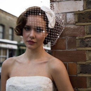 May include: A woman wearing a white bridal veil with a bow and pearl accents. She is wearing a white strapless dress with lace detailing. The background is a brick wall.