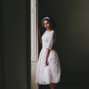 Puede incluir: Una mujer con un vestido blanco de encaje y velo se encuentra en una puerta. Lleva tacones negros y tiene el pelo recogido hacia atr&aacute;s.