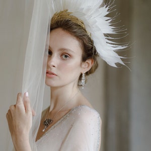 May include: A woman wearing a white feather crown with gold accents and a white dress with a beaded neckline. She is holding a sheer white fabric in front of her.