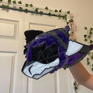 Peut inclure: Une pièce d'art artisanale, plate et bidimensionnelle en forme de tête d'animal stylisée. Le design présente une palette de couleurs violet foncé, noir et blanc, avec des contours noirs marqués définissant les traits, les oreilles et un bonnet.