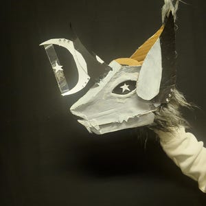 Puede incluir: Una máscara artesanal con diseño de lobo, hecha de cartón y pintada en blanco, negro y gris. Presenta un ojo en forma de estrella y un detalle de media luna. La máscara se sostiene sobre un fondo negro.