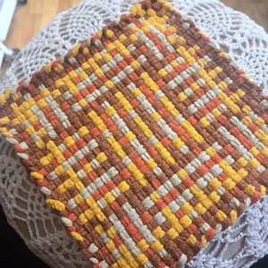 May include: A square potholder featuring a woven pattern of yarn in shades of brown, orange, yellow, and cream. The potholder is constructed with a dense, interlocking grid design and includes a small, circular yellow plastic loop at the top corner.