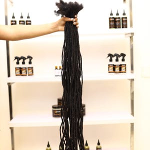 May include: A long set of black dreadlocks, held in a person's hand. The dreadlocks are about 2 feet long and are made of human hair.