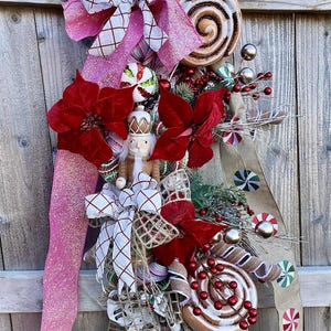 May include: A festive Christmas wreath with a wooden nutcracker, red poinsettias, and faux cinnamon rolls. The wreath is adorned with pink glitter ribbon, white and red patterned bows, and candy-themed ornaments.