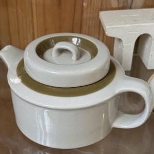 Puede incluir: Una tetera de cerámica color crema con cuerpo redondeado, asa curva y boquilla. La tapa también es color crema con una banda marrón y un asa pequeña. La tetera forma parte de un juego de té.