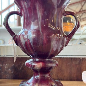 Op de afbeelding: Een bordeauxrode keramische vaas met twee gebogen handvatten. De vaas heeft een gegroefd ontwerp en staat op een vierkante basis met een turquoise en bordeauxrode glazuur. De vaas is een decoratief item.