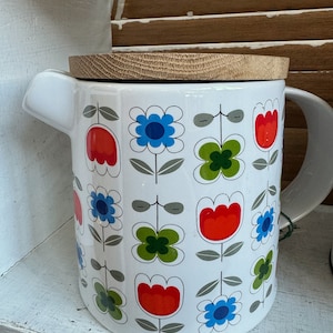 Puede incluir: Jarra de cerámica blanca con tapa de madera. La jarra está decorada con un patrón repetido de flores coloridas en rojo, azul y verde. Tiene un asa grande y un pico para verter.