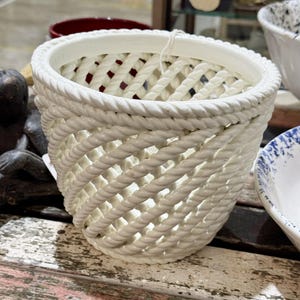 May include: A white ceramic planter featuring a decorative lattice design that mimics the appearance of twisted rope. The vessel has a wide, circular opening and a tapered base, with a smooth, glossy finish throughout the entire exterior surface.