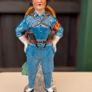 May include: A ceramic figurine of a woman wearing a blue uniform with a red star on her sleeve. She is holding a rifle and has a basket on her back. The figurine is standing on a base with a green leaf design.