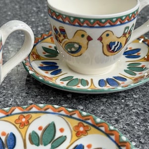 Puede incluir: Juego de tazas y platillos de cerámica con un diseño de pájaros coloridos. Las tazas y los platillos tienen una base blanca con detalles amarillos, azules y verdes. Las tazas tienen asa y los platillos tienen un borde decorativo.