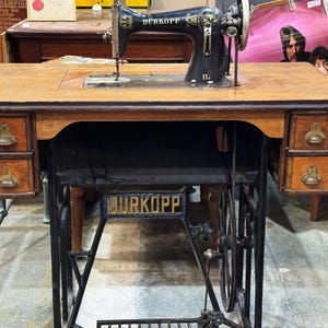 Pode incluir: Uma antiga máquina de costura Dürkopp com uma mesa de madeira e duas gavetas de cada lado. A máquina é preta com detalhes dourados e assenta numa estrutura de metal preta. A mesa é feita de madeira castanha clara.