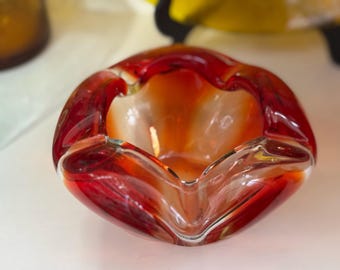 May include: A handcrafted ashtray made of red and orange glass. The ashtray has a unique, organic shape with a clear, smooth interior. The glass is translucent, allowing light to pass through, and has a glossy finish.