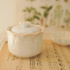 May include: White ceramic jar with a lid, featuring a floral design. The jar is sitting on a sheet of music.