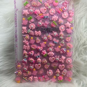 May include: A clear phone case adorned with numerous pink charms, including flowers, hearts, stars, and small figures. The word "Happy" is visible, along with the name "Zaynah". A light pink ribbon is tied at the top.