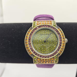 Puede incluir: Un reloj de pulsera dorado con una correa de ante morado. La esfera y el bisel del reloj están incrustados con pequeños cristales de colores en tonos verdes, naranjas y morados. El reloj se muestra sobre una superficie negra.