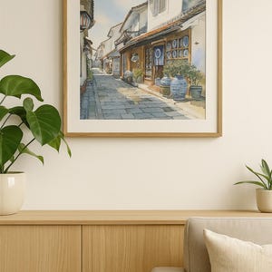 Puede incluir: Pintura a la acuarela de una calle estrecha con edificios tradicionales y un camino empedrado. Los edificios tienen fachadas de madera y tejados de tejas. Hay macetas de cerámica azul y blanca con plantas frente a los edificios. La obra está en un marco de madera.