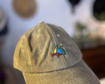 May include: A khaki-colored baseball cap with a curved brim. The cap features a small, colorful embroidered design of a helicopter on the front. The cap has a classic six-panel design with a button on top.