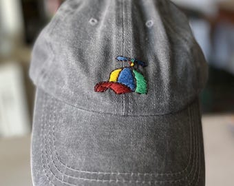 May include: A charcoal gray baseball cap featuring an embroidered design on the front panel. The embroidery depicts a small, multi-colored propeller hat in shades of red, yellow, blue, and green, complete with a tiny propeller on top.